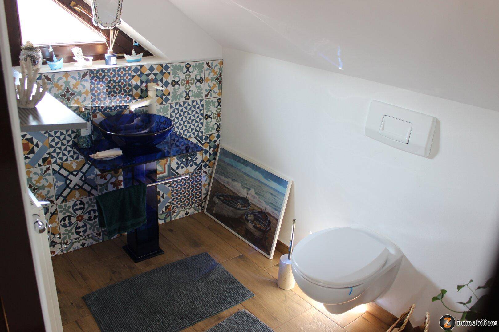 Kleines Badezimmer im Dachgeschoss mit Wand-WC, modernem Waschbecken und bunten Fliesen.