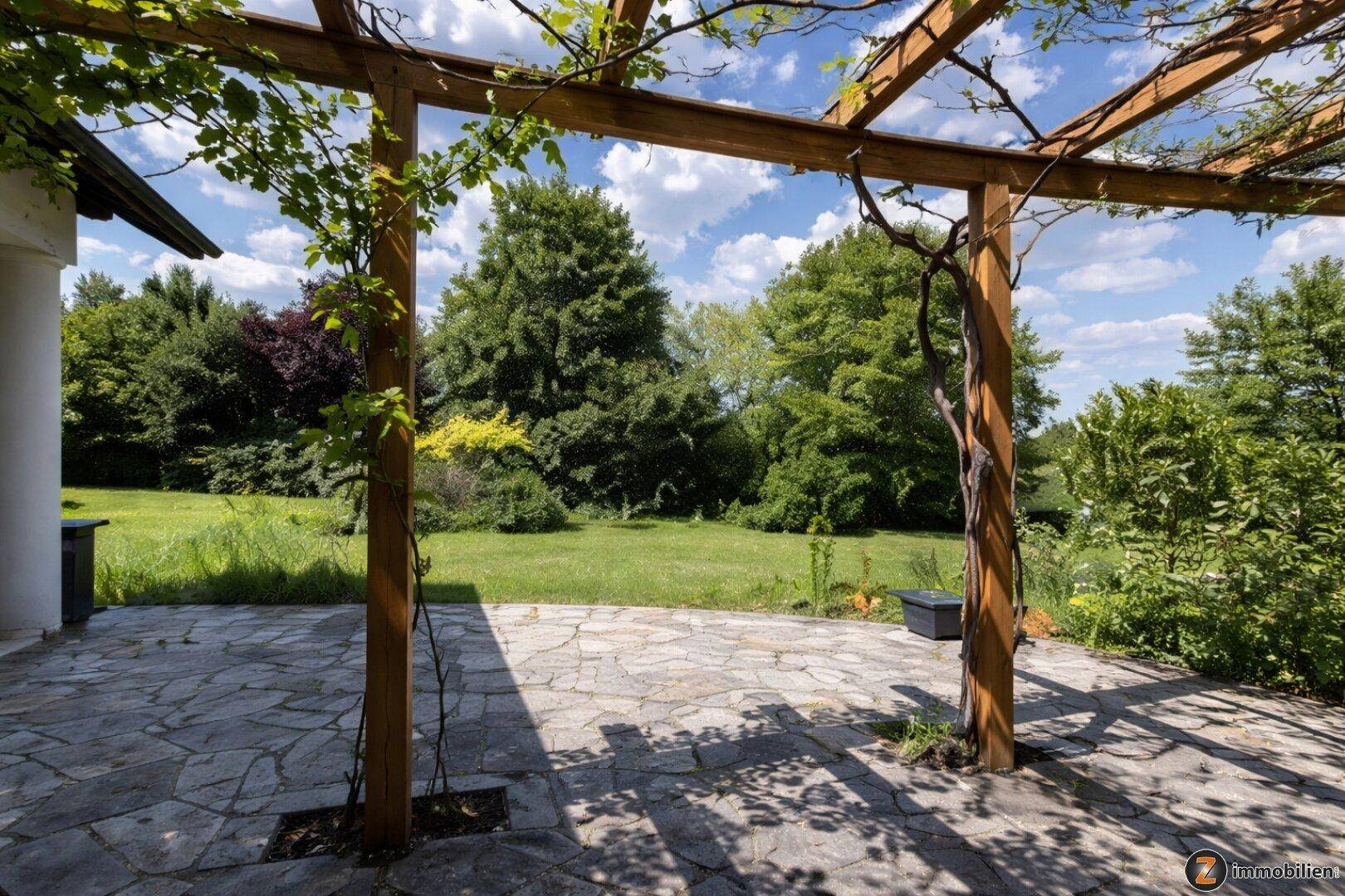 Steinplatten-Terrasse mit Holzpergola und üppiger Begrünung, umgeben von einem großen Garten.