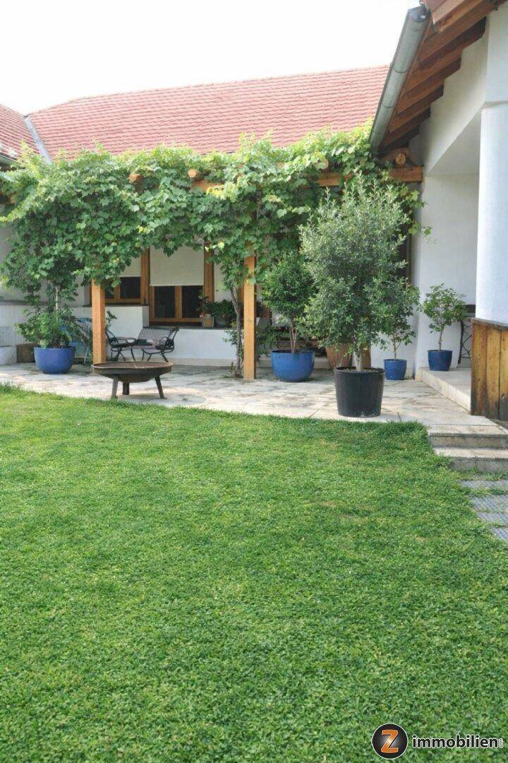 Grüner Garten mit Rasenfläche und Zugang zu einer überdachten Terrasse, umgeben von Pflanzen.