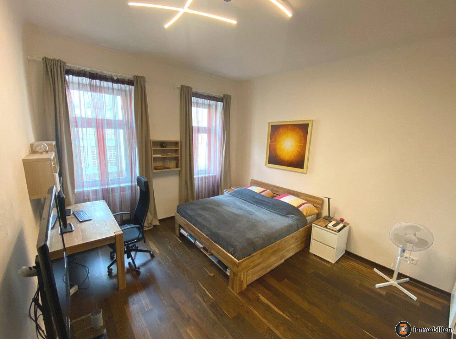 Helles Schlafzimmer mit Doppelbett, Schreibtisch und dunklem Holzboden, ideal als Schlaf- und Arbeitsbereich.