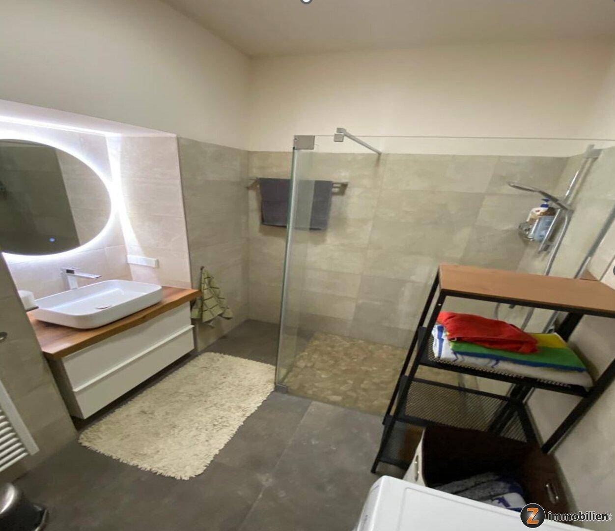 Elegantes Badezimmer mit modernem Waschtisch, beleuchtetem Spiegel und geräumiger begehbarer Dusche.