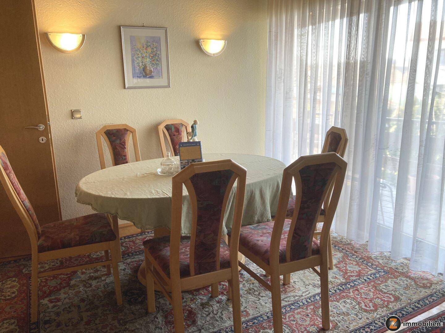 Essbereich mit rundem Tisch, Holzstühlen und Teppichboden neben einem großen Fenster.