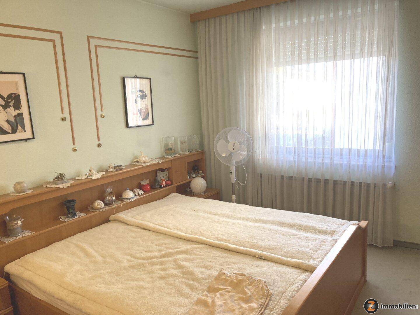 Helles Schlafzimmer mit Doppelbett, Fenster und Heizkörper, ausgestattet mit einem Ventilator.