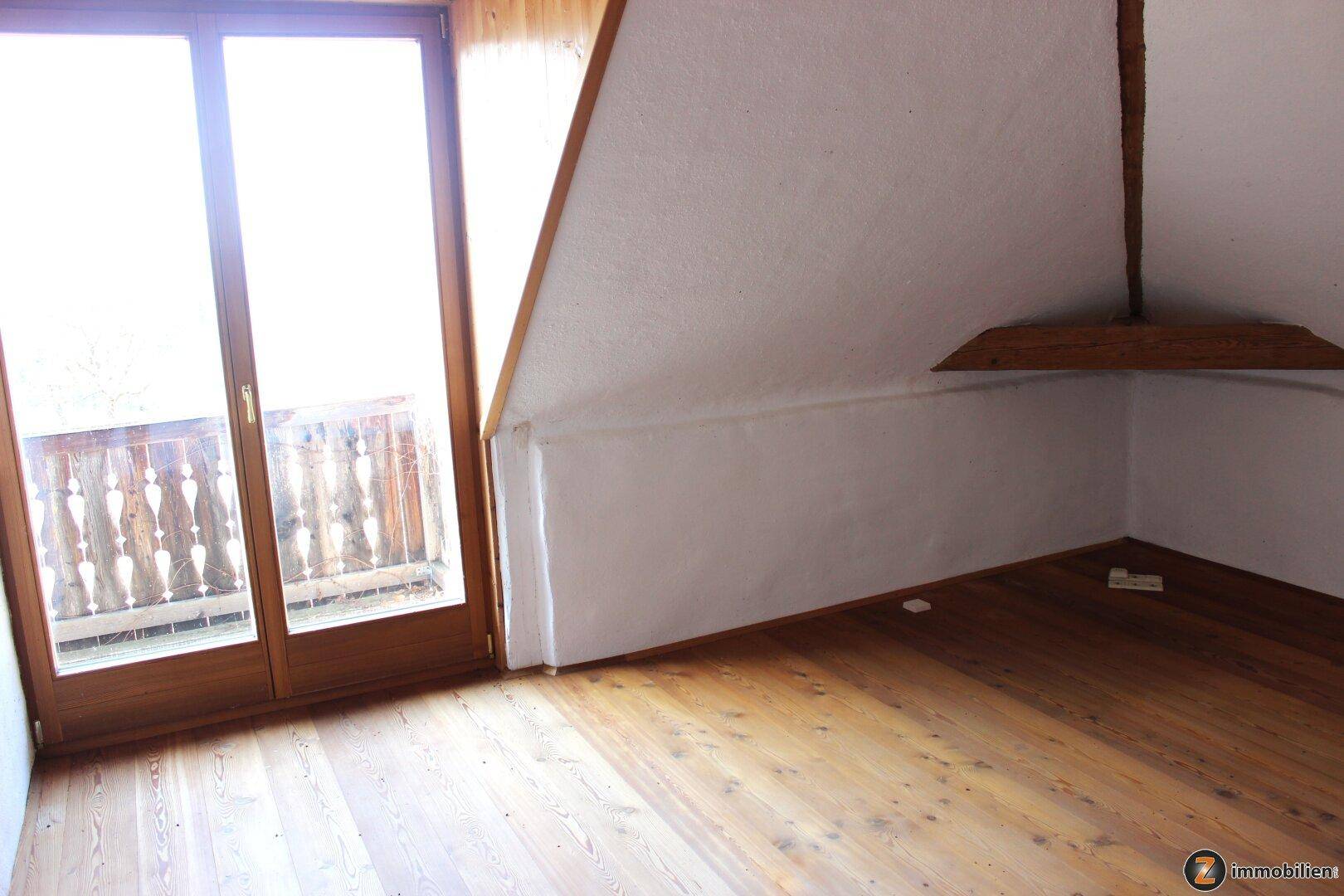 Helles Zimmer mit Holzboden, Dachschräge und Zugang zu einem Balkon.