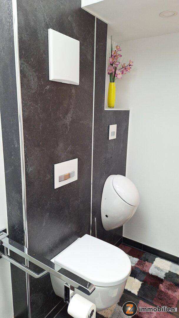 Separate Toilette mit Wand-WC, Urinal und dunklen Wandfliesen, dekoriert mit Blumen.