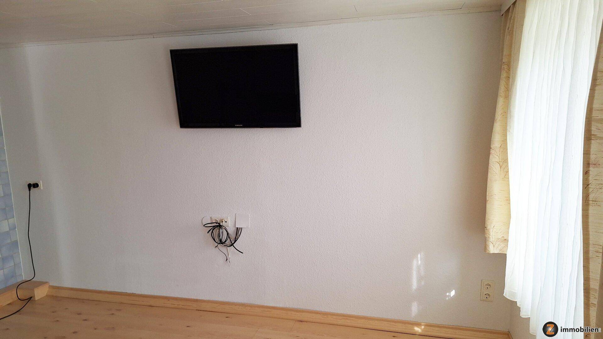 Weiße Wand mit montiertem Fernseher und sichtbaren Kabeln, daneben ein Fenster mit Vorhang.
