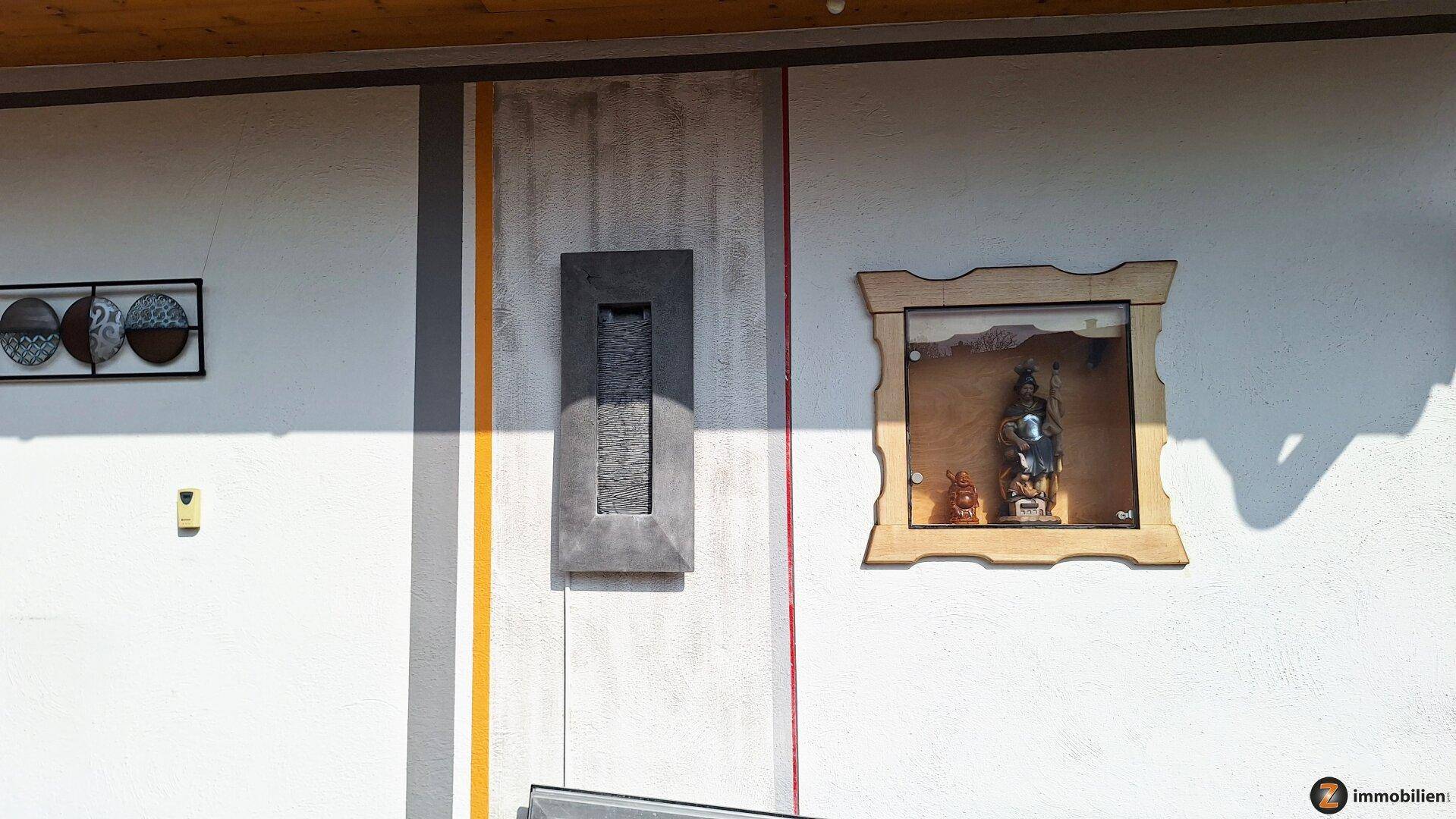 Dekorative Elemente an der Außenwand des Hauses, darunter ein Wandbrunnen und ein Holzrahmen.