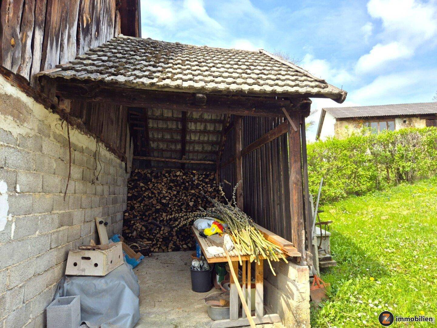 Offener Holzschuppen mit gelagertem Brennholz und Blick auf die grüne Umgebung.