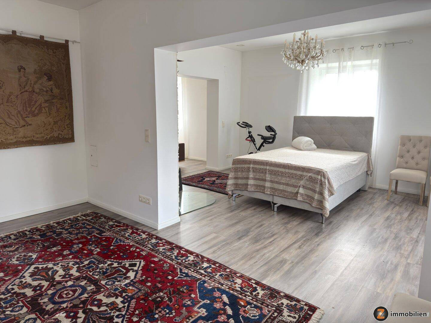 Helles Schlafzimmer mit großem Bett, Trainingsgerät und Fenster mit Vorhängen.