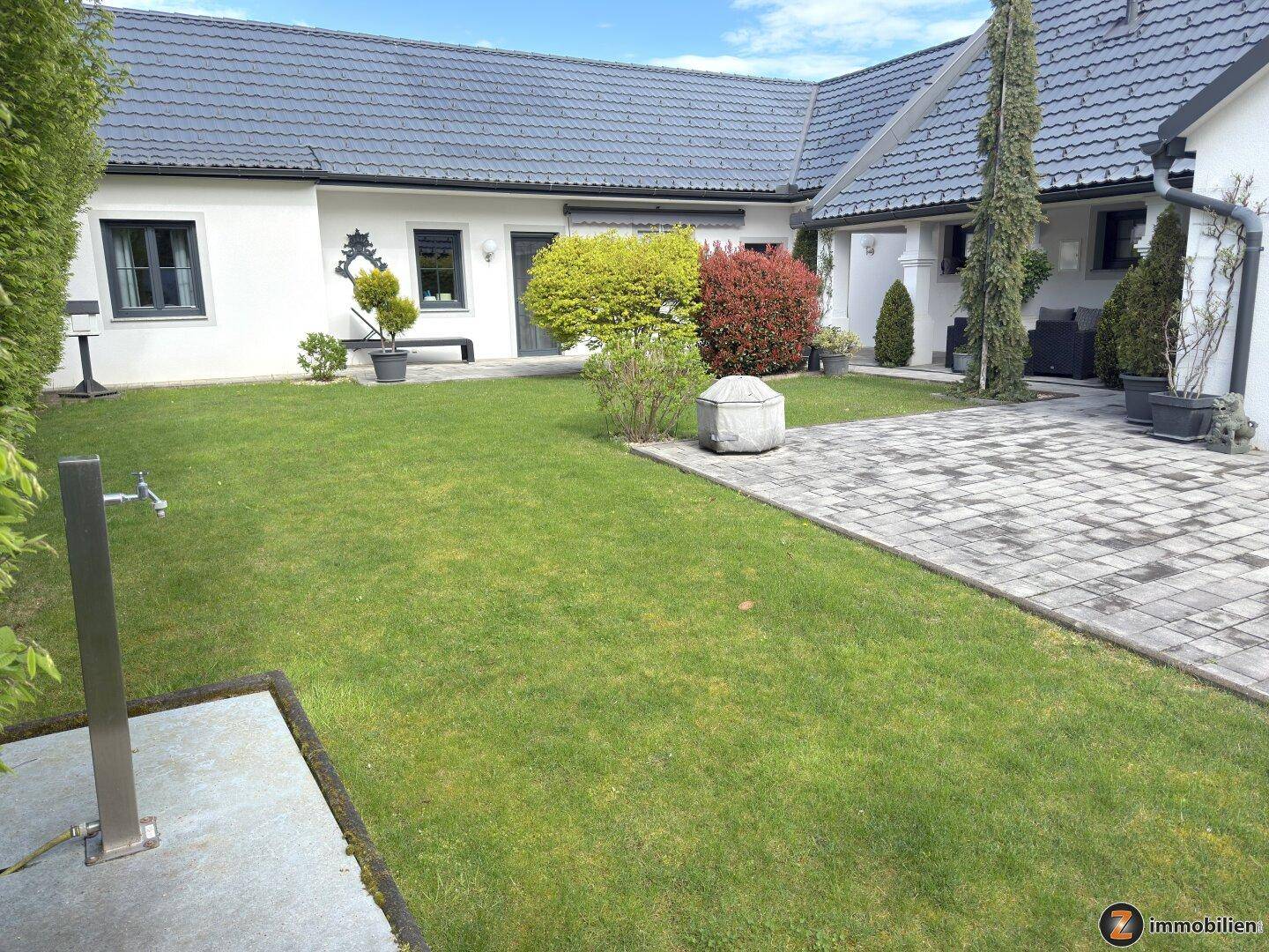 Gepflegte Rasenfläche mit gepflastertem Weg und Außendusche im sonnigen Gartenbereich.