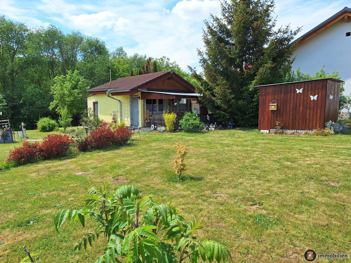 Kleines Haus mit Holzschuppen und weitläufigem Garten unter einem sonnigen Himmel.