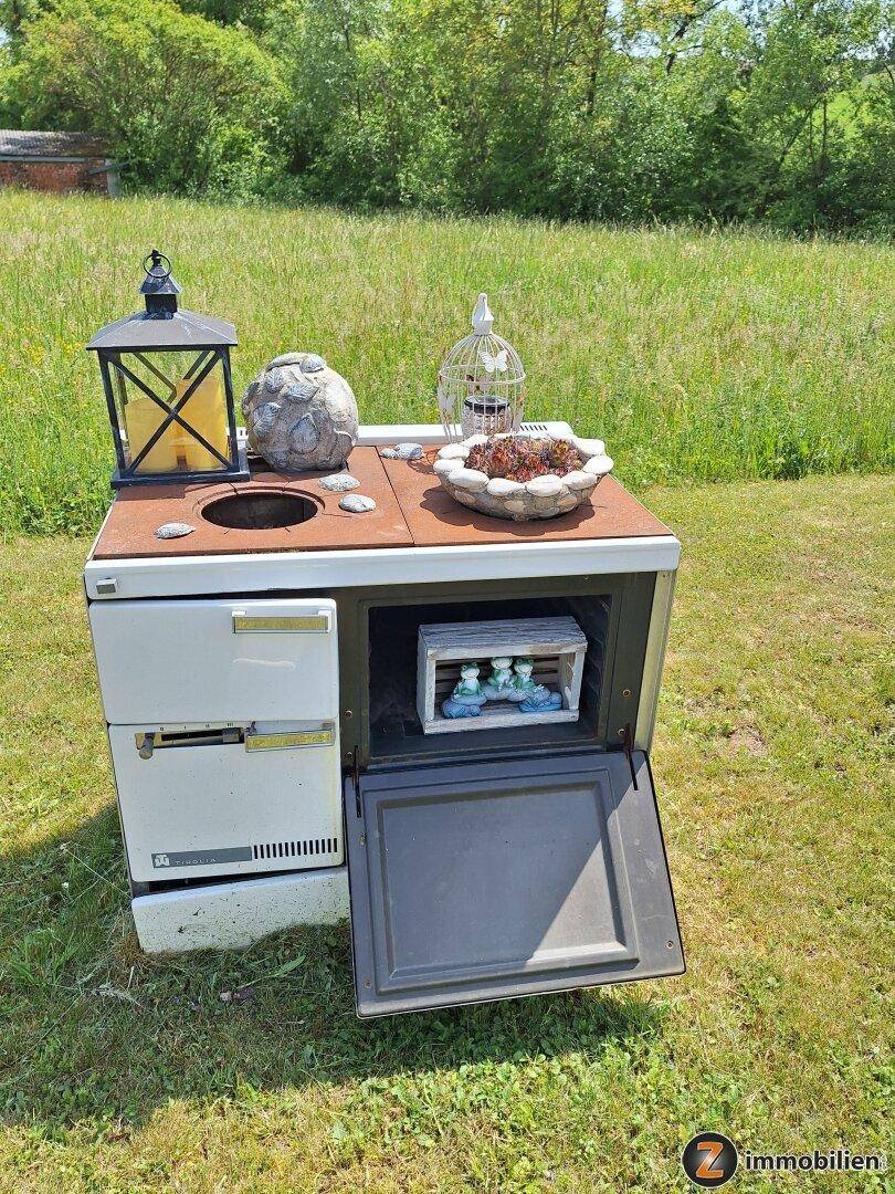 Dekorativer alter Ofen im Garten, umgeben von Grünflächen und ländlicher Umgebung.