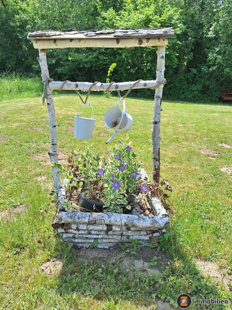 Rustikaler Zierbrunnen aus Birkenholz mit Blumen im Garten, umgeben von Rasen.