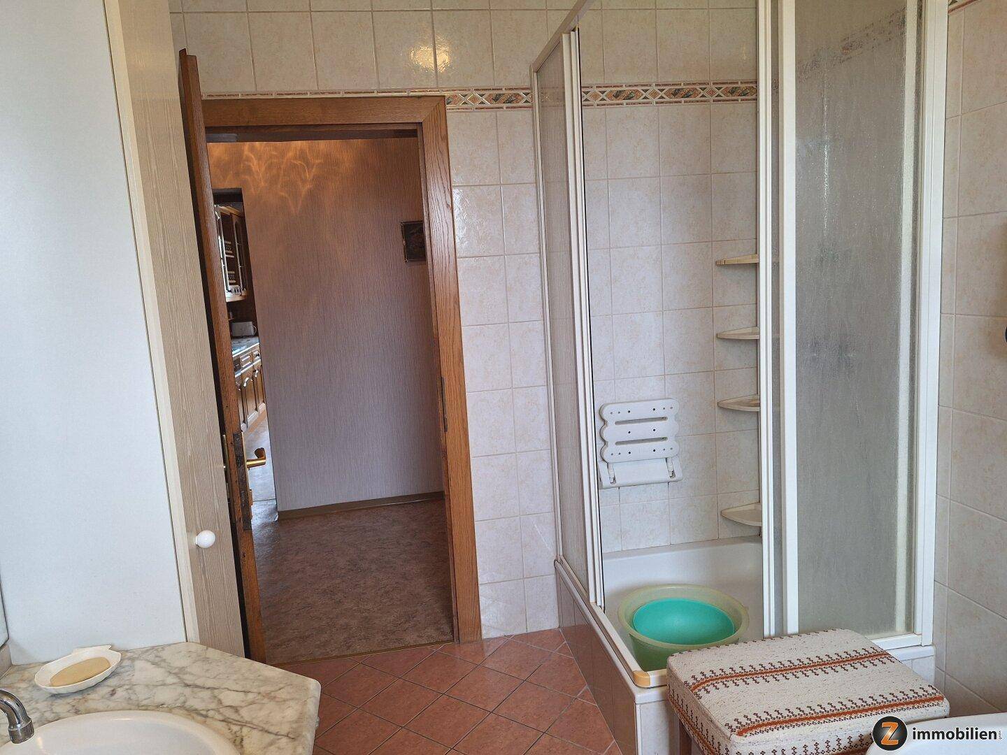 Badezimmer mit Duschkabine, Waschbecken und gefliesten Wänden, mit Zugang zu einem anderen Raum.