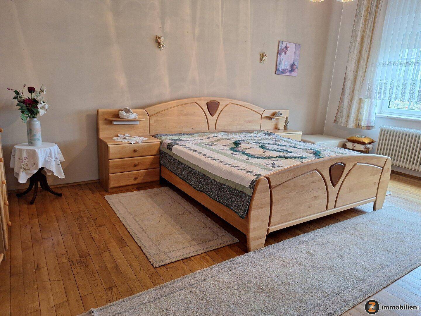 Geräumiges Schlafzimmer mit hellem Holzbett, Nachttischen und Teppichen auf Holzboden.