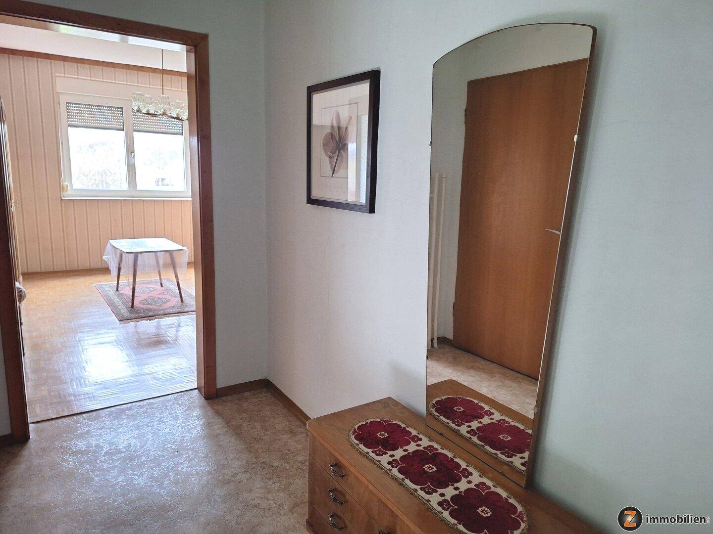 Flur mit großem Spiegel, Kommode und Zugang zu einem hellen Zimmer mit Fenster.