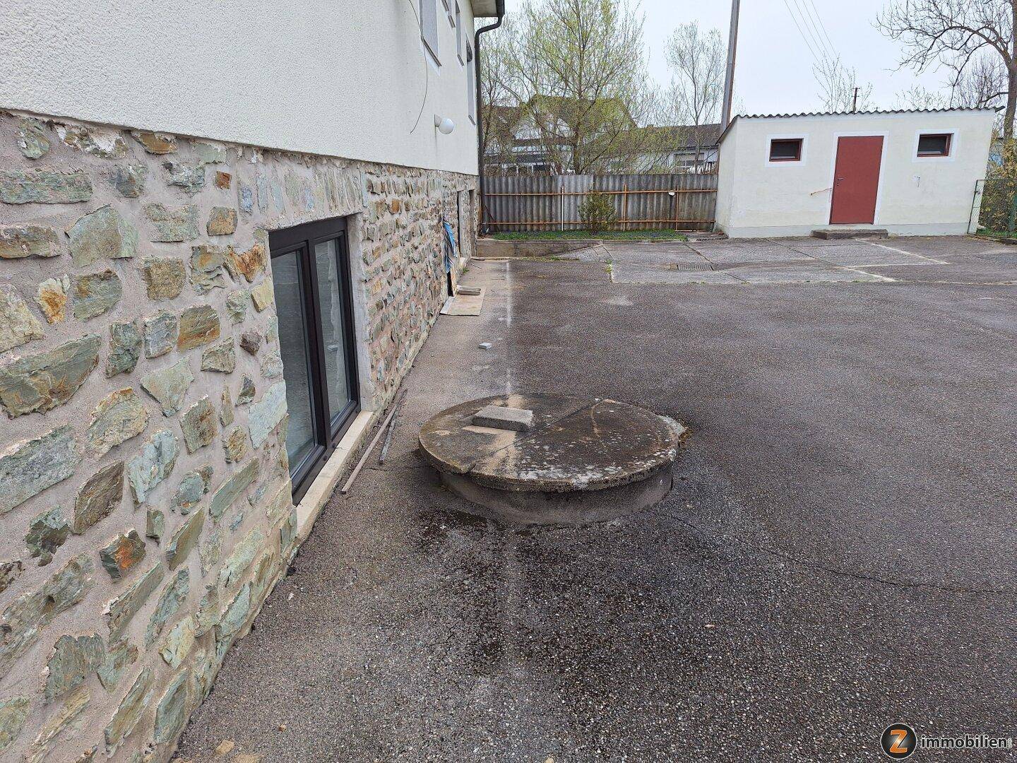 Seitenansicht des Gebäudes mit Steinfassade, Fenster und einem Schuppen im Hintergrund auf asphaltiertem Gelände.