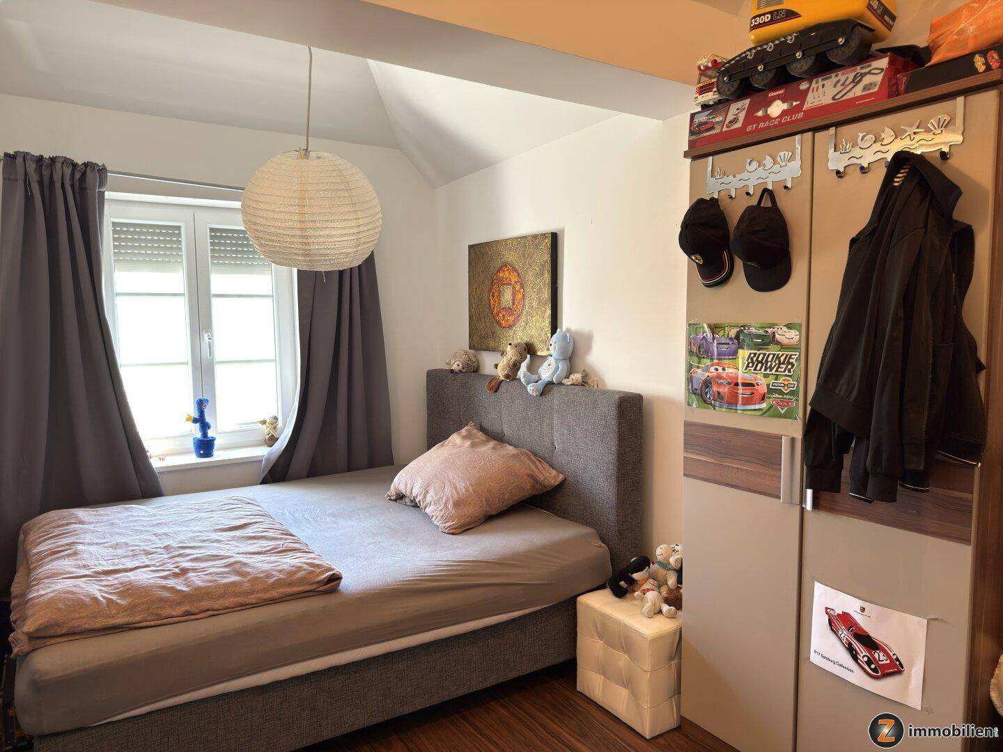 Kinderzimmer mit Einzelbett, Kleiderschrank und Fenster mit Vorhängen.
