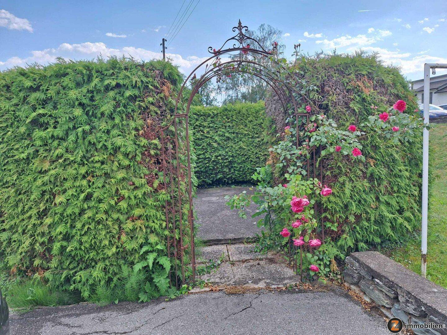 Ein schmiedeeisernes Tor, umrankt von blühenden Rosen, führt durch eine dichte Hecke in den Gartenbereich.