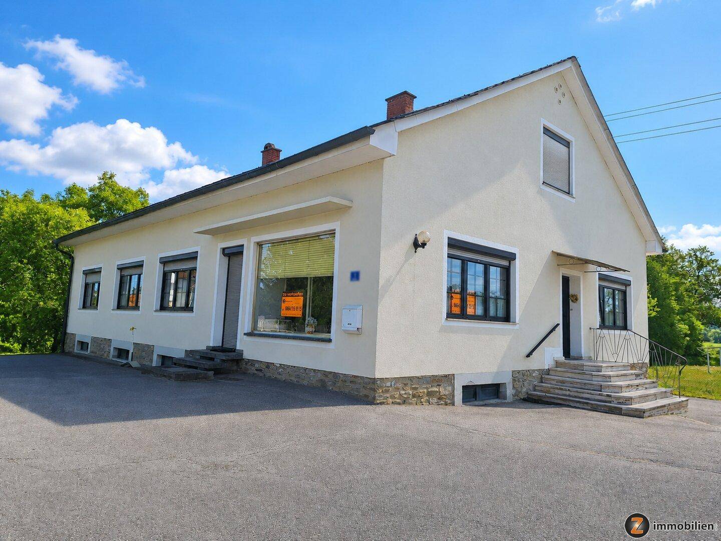Vorderansicht eines hellen Gebäudes mit Satteldach, mehreren Fenstern und einem Eingangsbereich mit Treppe.