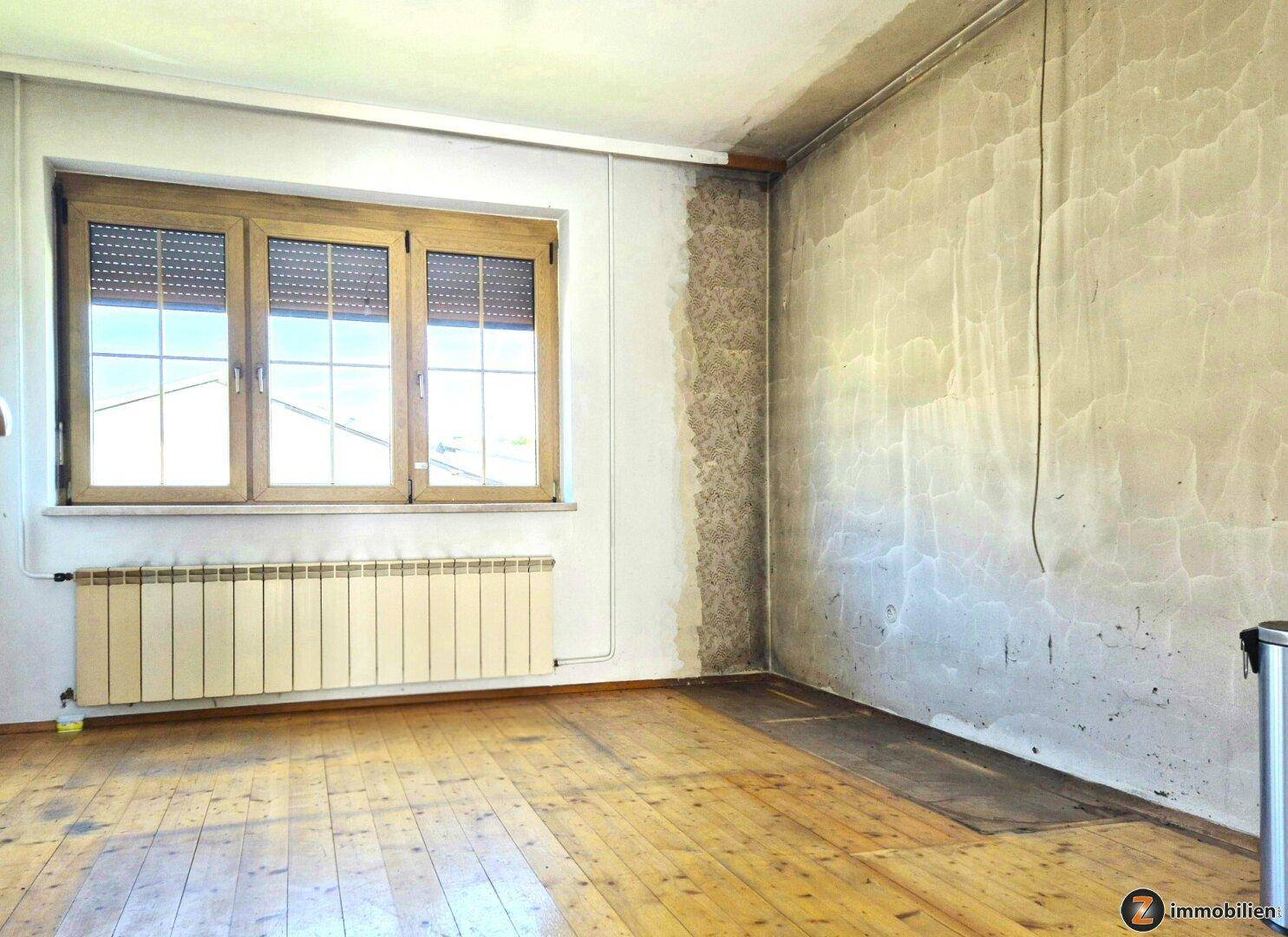 Leerer Raum mit Dielenboden, Fenster mit Rollläden und Heizkörper, die Wände sind renovierungsbedürftig.