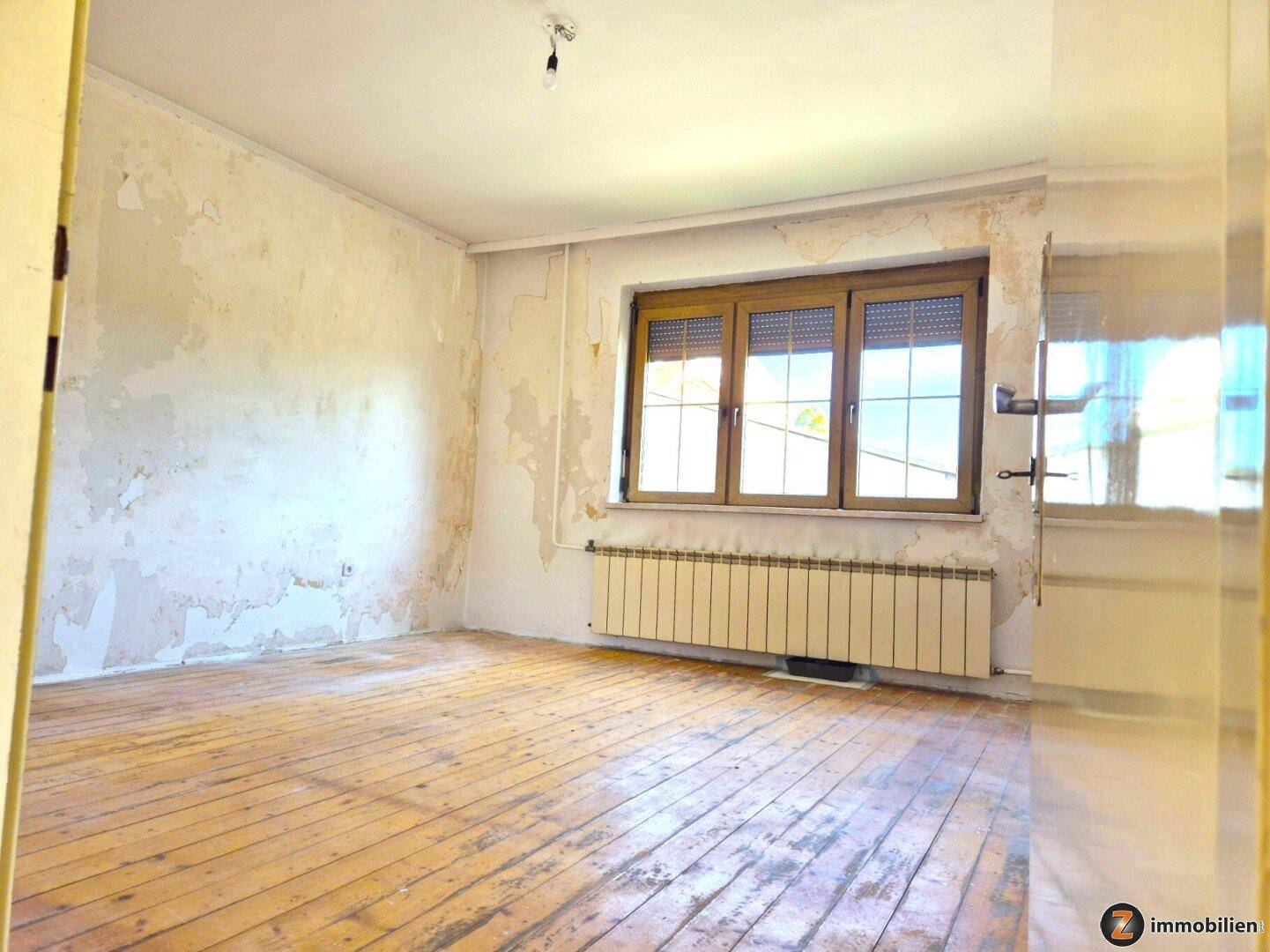Heller Raum mit Holzdielenboden und Fenster mit Rollläden, die Wände zeigen deutliche Abnutzungsspuren.