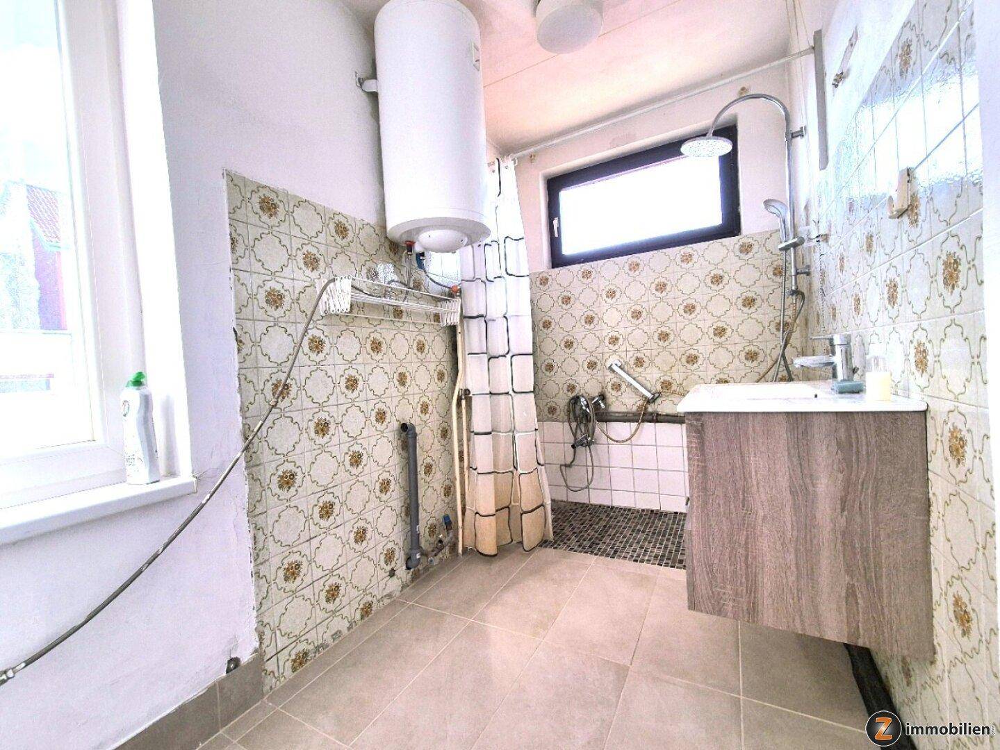 Badezimmer mit gemusterten Fliesen, Duschbereich, Waschbeckenunterschrank und Warmwasserboiler.