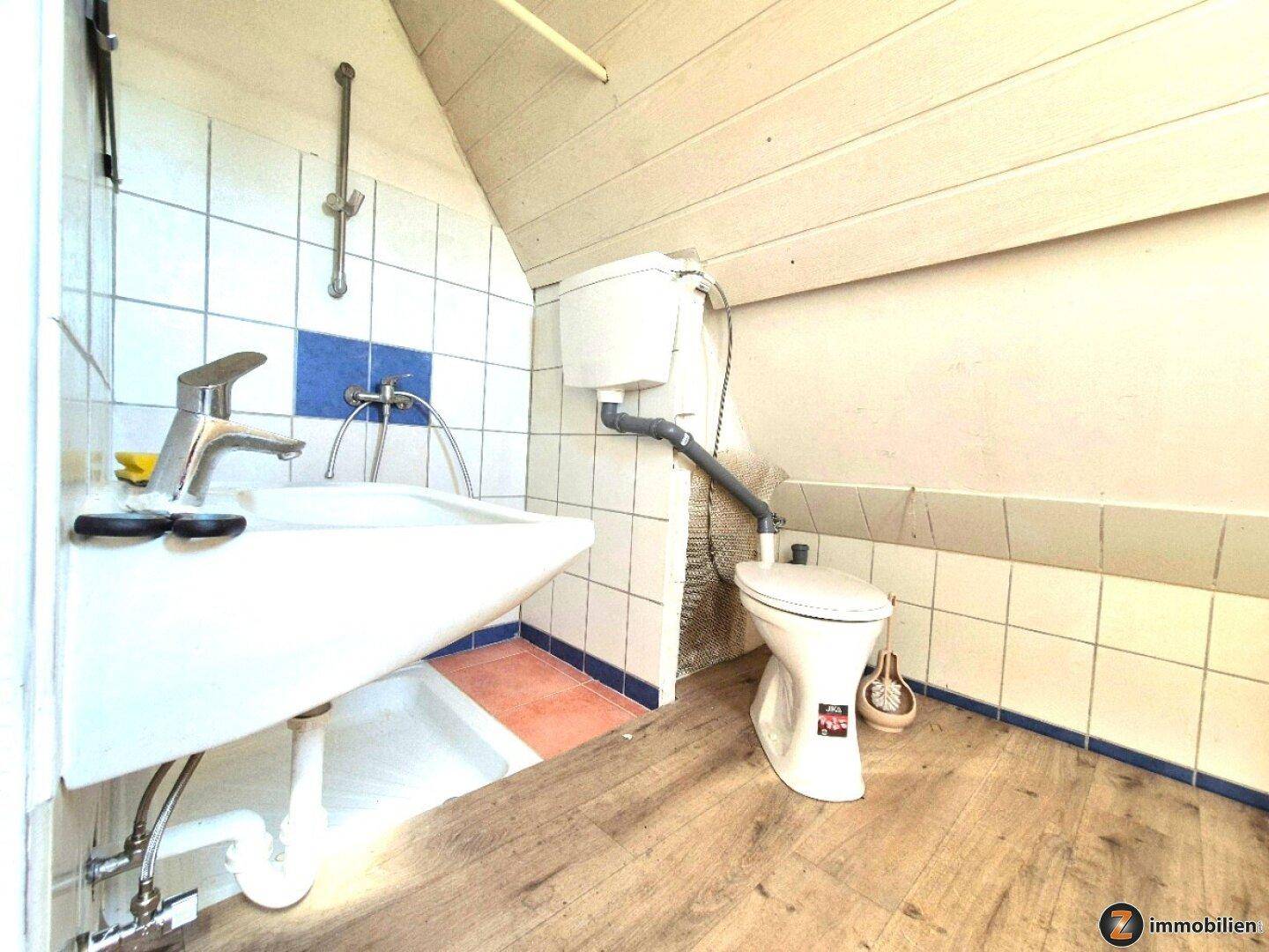 Kleines Badezimmer mit WC, Waschbecken und Duschvorrichtung unter einer Dachschräge.