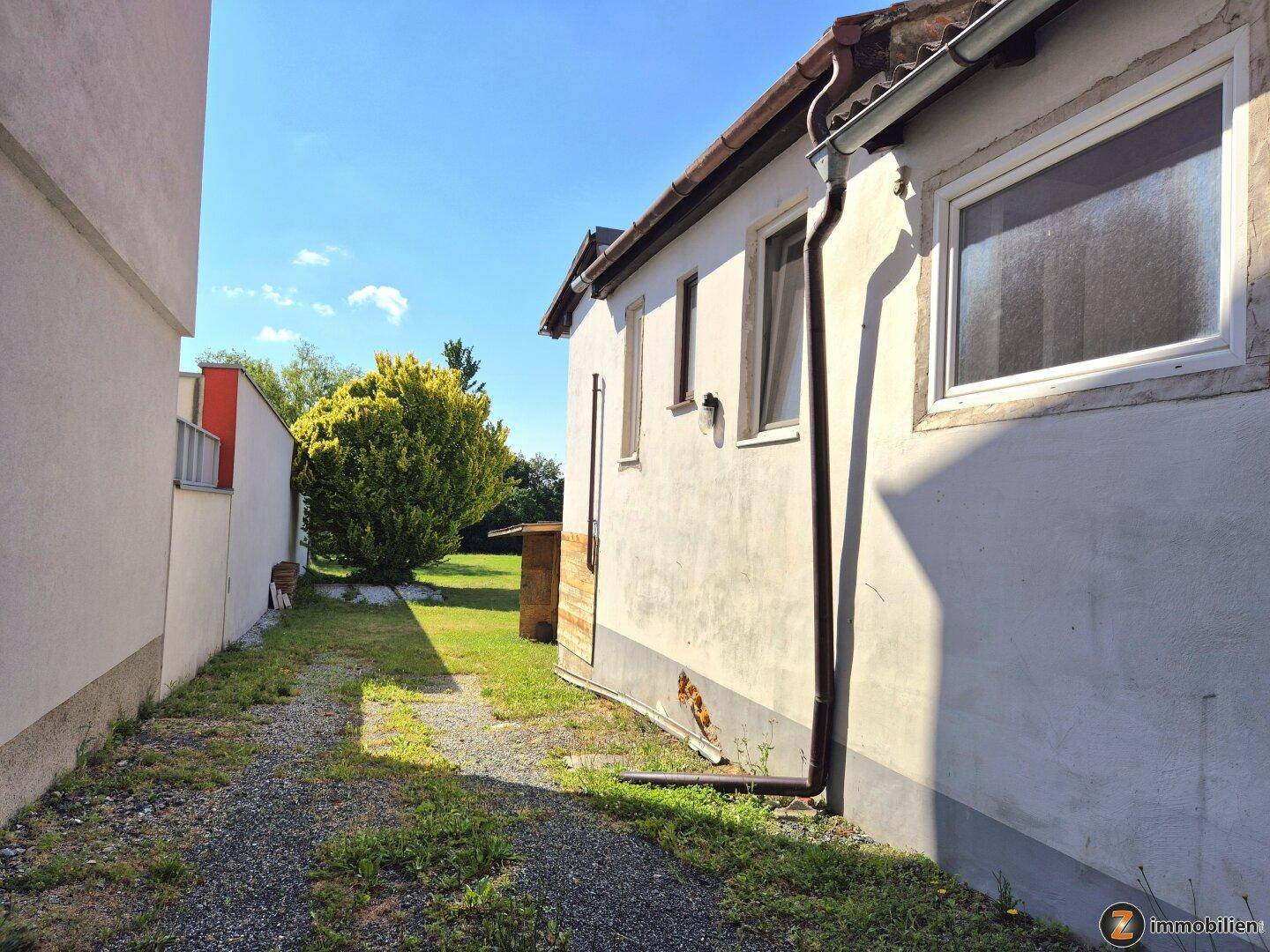 Seitenweg neben dem Haus mit Kies und Gras, der zu einer grünen Fläche führt.