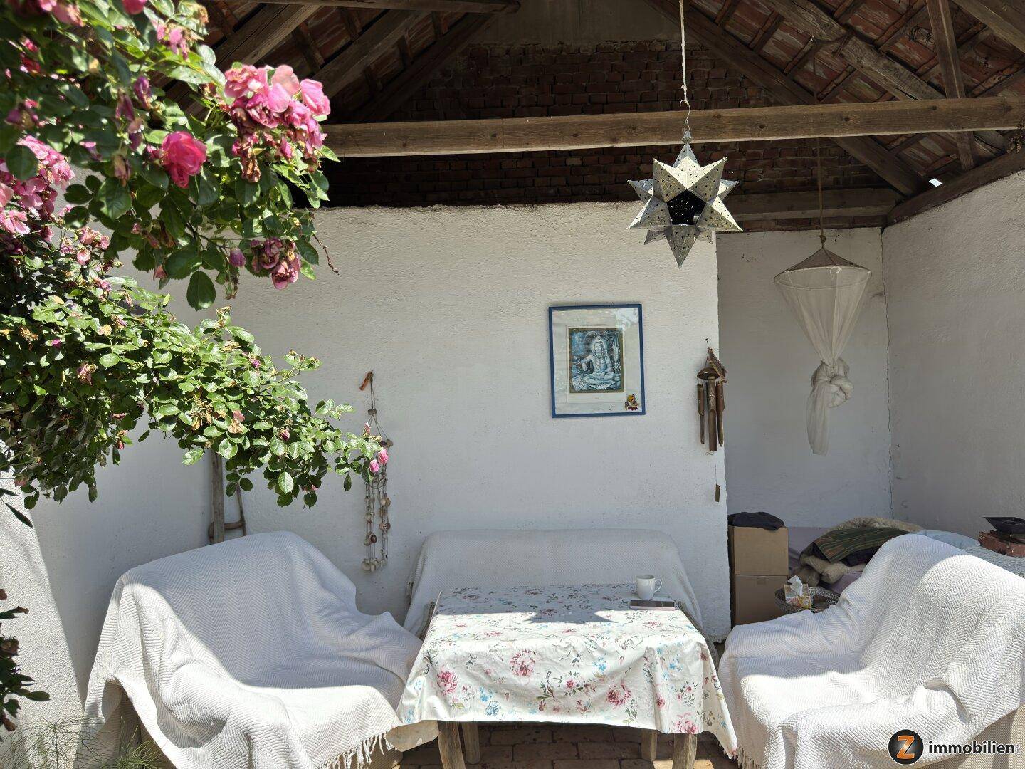 Überdachte Terrasse mit Gartenmöbeln, umgeben von blühenden Pflanzen und viel Grün.