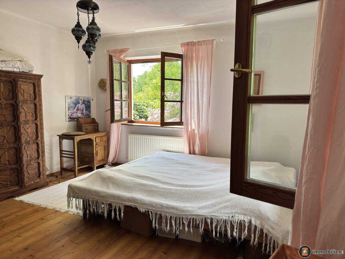 Schlafzimmer mit Holzboden, einem großen Bett und einem Fenster mit Blick ins Grüne.