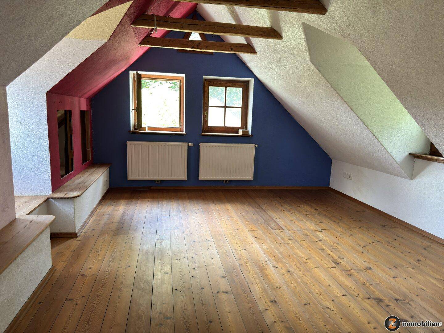 Geräumiger Dachbodenraum mit Holzboden, farbigen Wänden und zwei Fenstern.