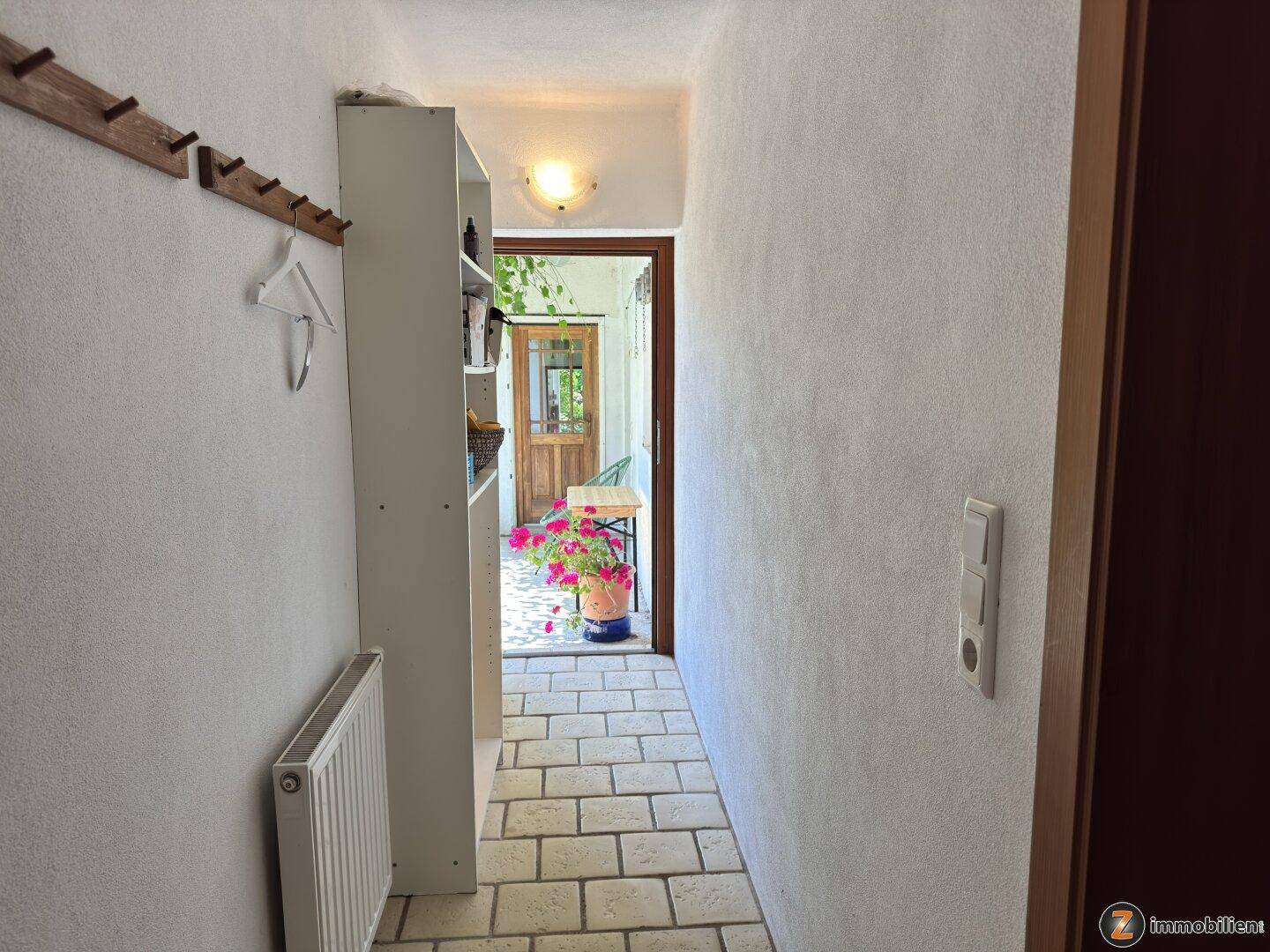 Schmaler Flur mit Fliesenboden, Garderobe und Blick auf einen sonnigen Außenbereich.
