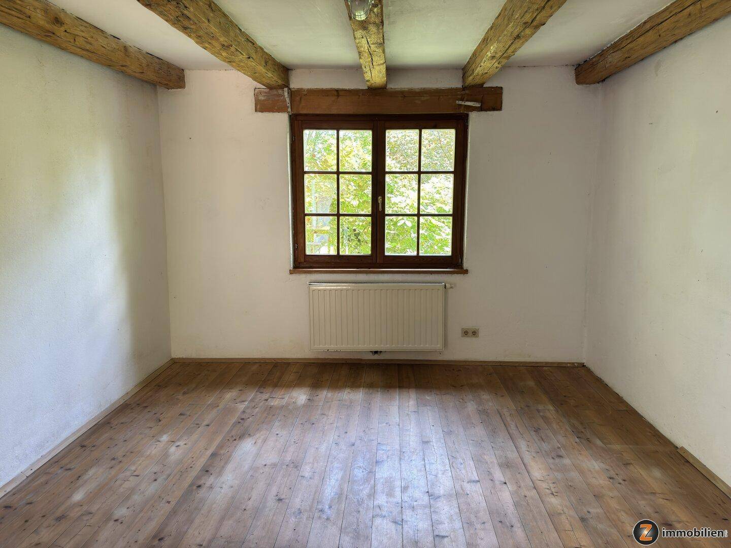 Einfacher Raum mit Holzbalken, Holzboden und einem großen Fenster mit Blick ins Grüne.