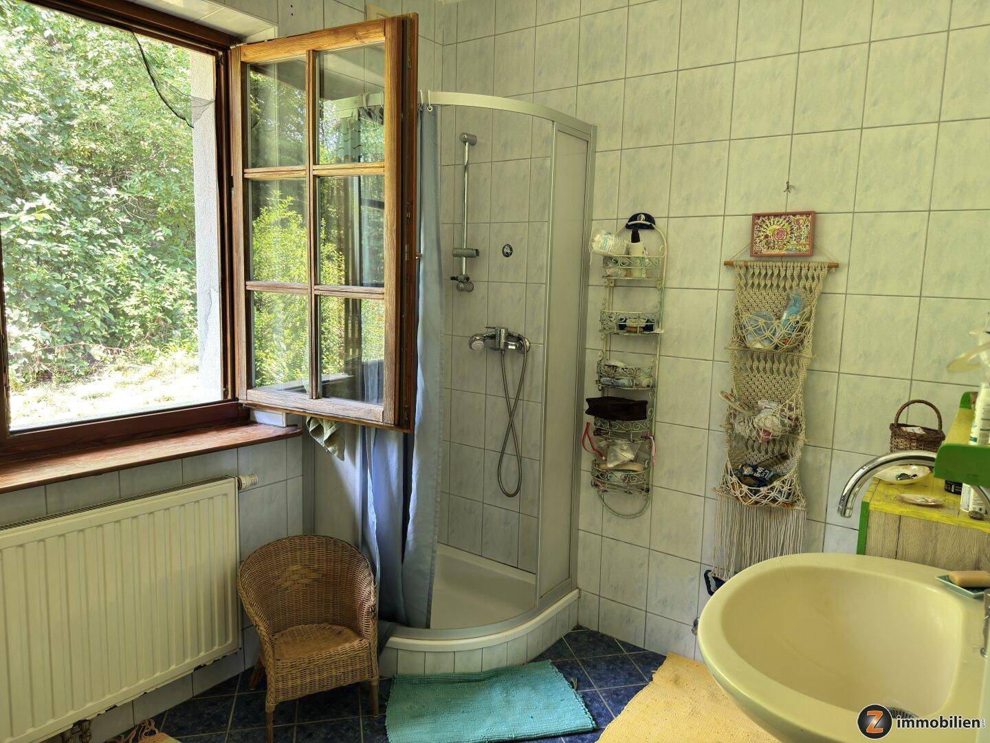 Badezimmer mit blauen Fliesen, Duschkabine und einem Fenster mit Blick ins Grüne.