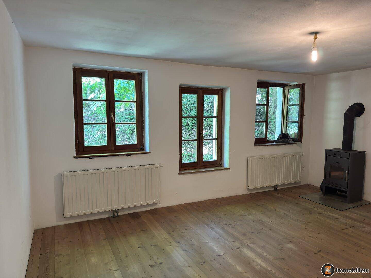 Geräumiges Wohnzimmer mit mehreren Fenstern, Holzboden und einem gemütlichen Kaminofen.