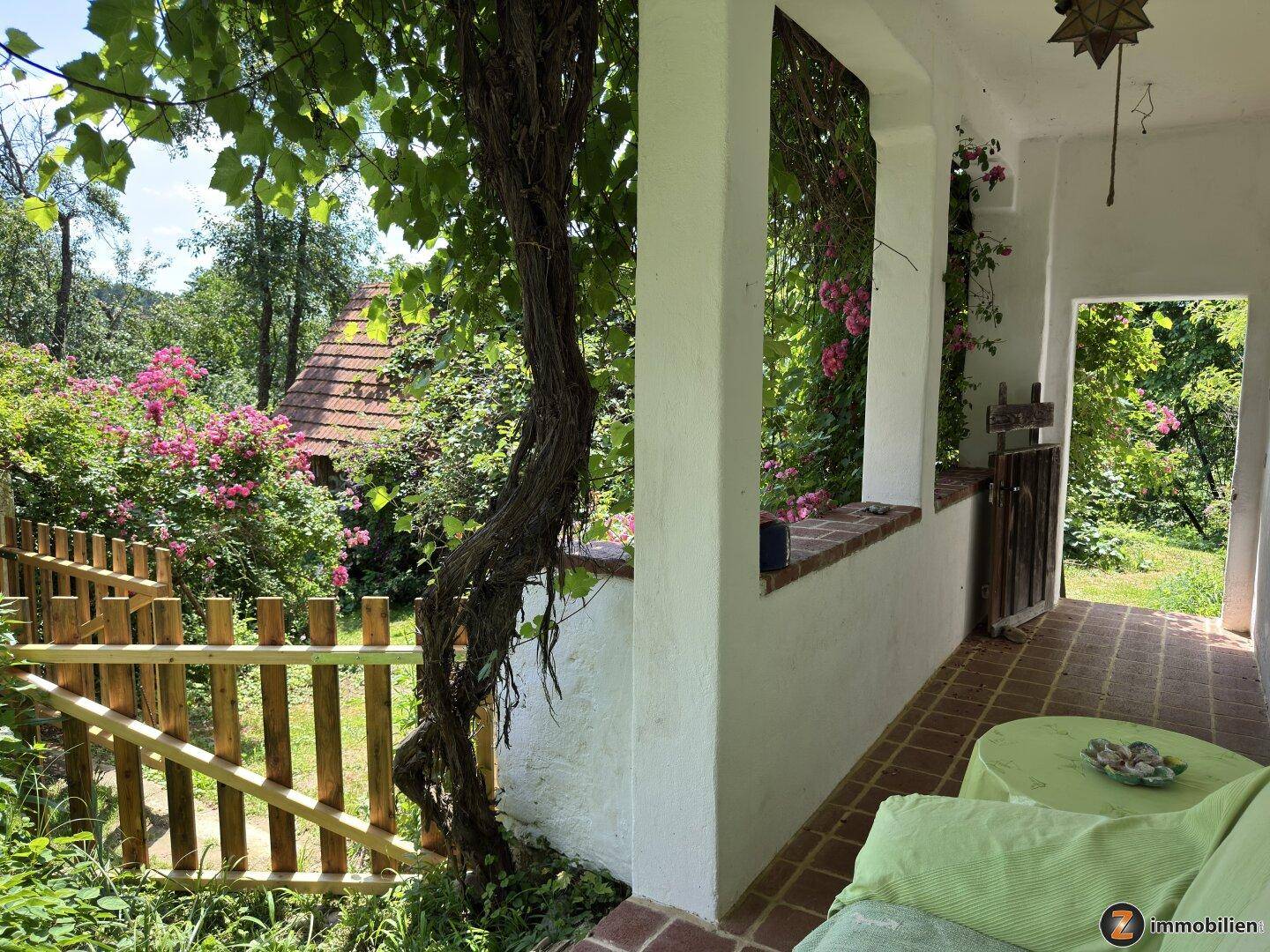 Überdachter Außenbereich mit Sitzgelegenheit, umgeben von üppiger Vegetation und einem Holzzaun.