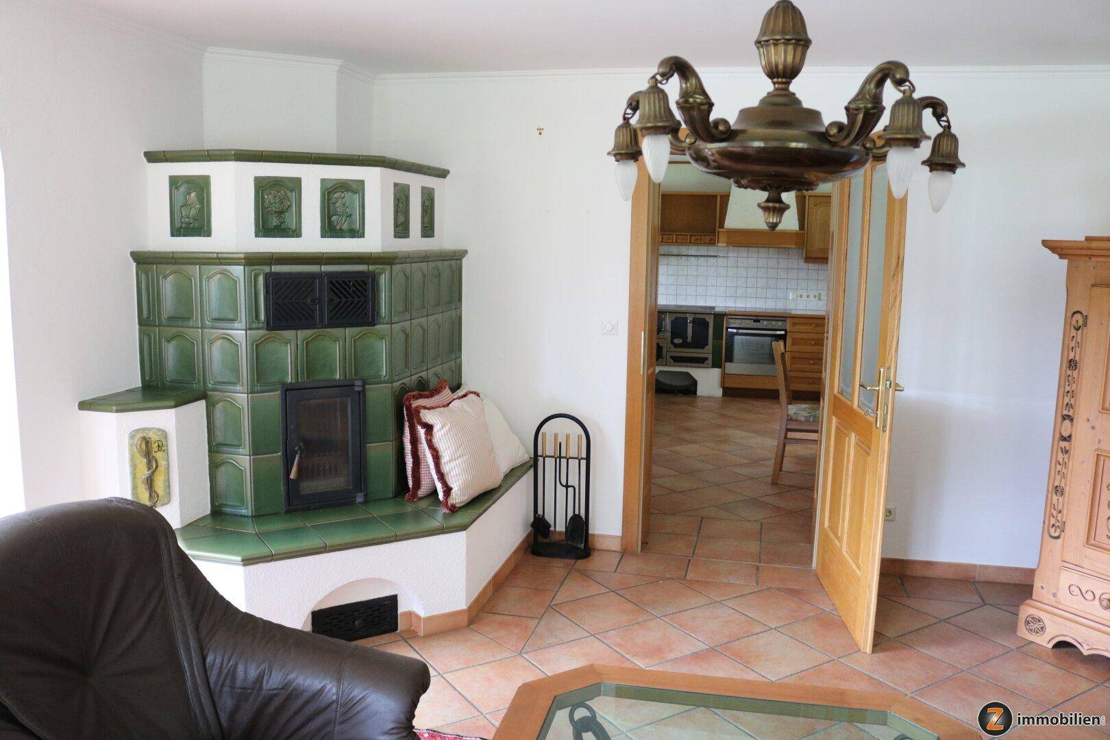 Gemütliches Wohnzimmer mit grünem Kachelofen, Terrakottafliesen und Zugang zur Küche.