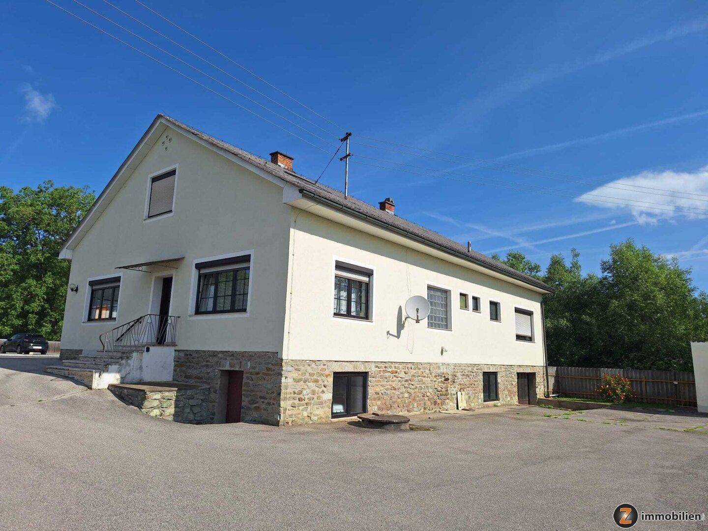 Einfamilienhaus mit heller Fassade, Steinsockel und Satteldach, umgeben von Grünflächen und einem Vorplatz.