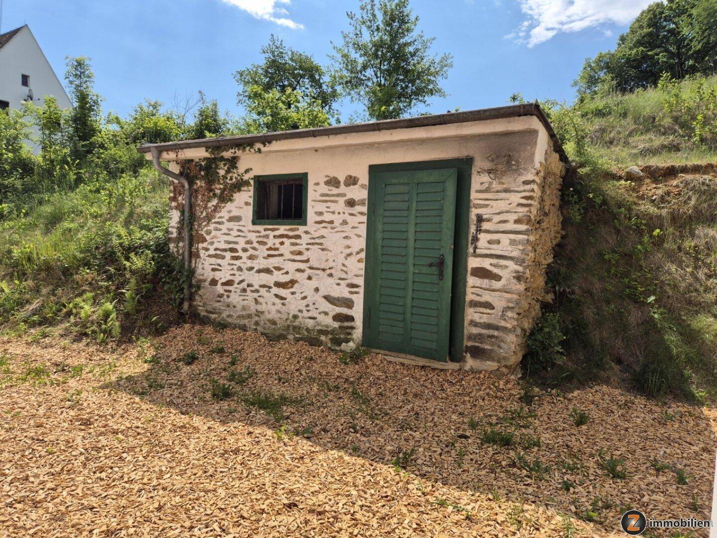 Rustikaler Steinschuppen mit grüner Holztür und Fenster, eingebettet in die Natur.