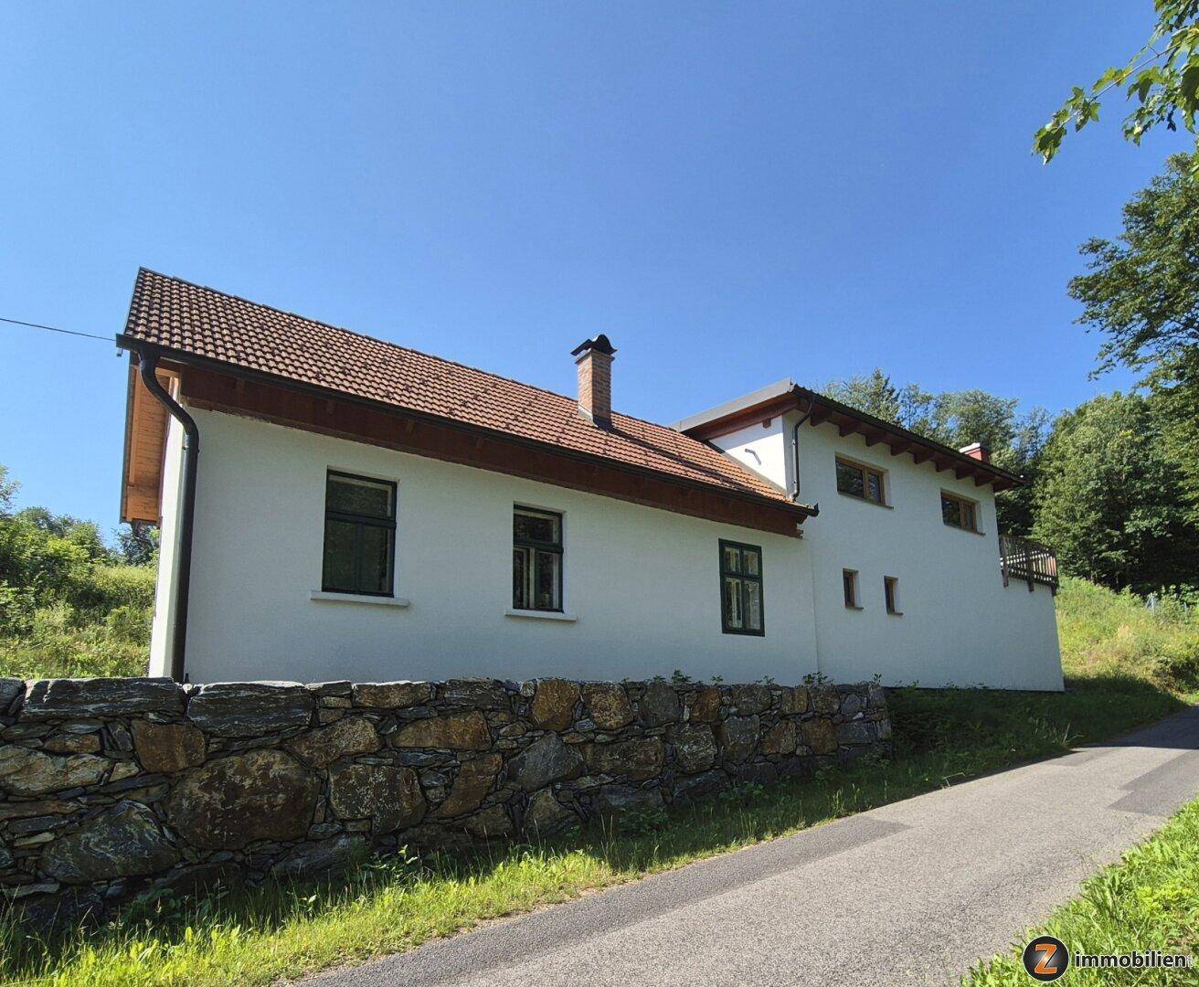 Rückansicht des Hauses mit weißer Fassade, Ziegeldach und Steinmauer im Vordergrund.