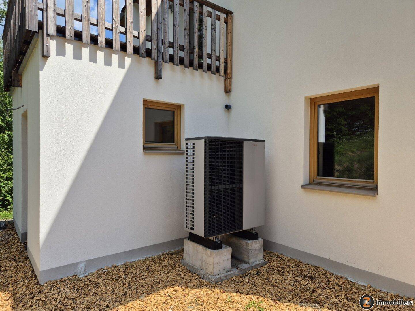Moderne Wärmepumpe an der Hauswand, umgeben von Holzhackschnitzeln und Natur.