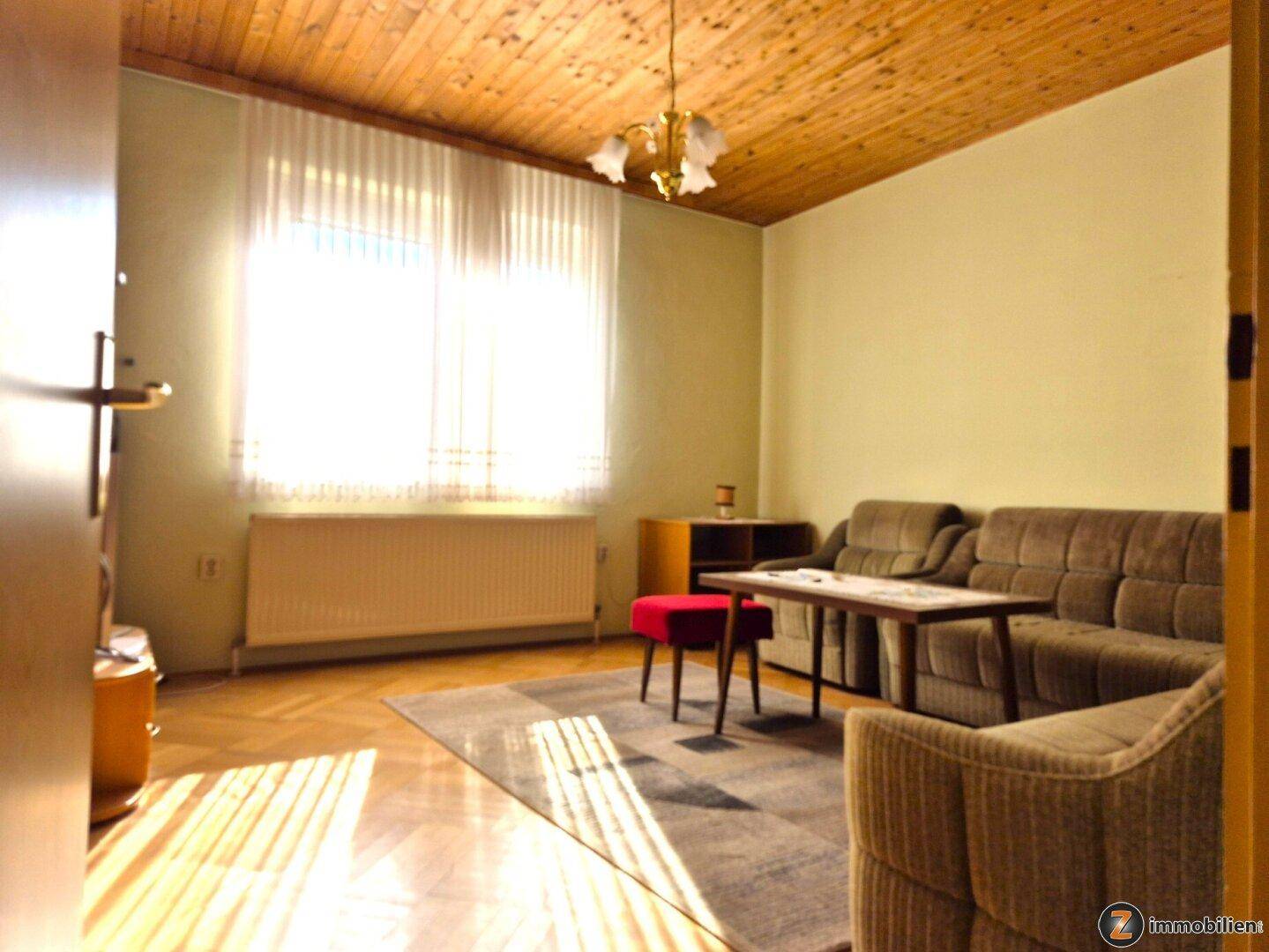 Wohnzimmer mit Holzdecke, Sofa und Teppich, hell durch großes Fenster.