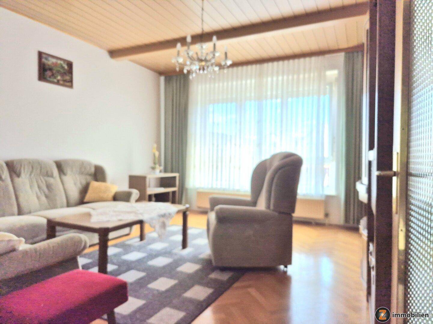 Gemütliches Wohnzimmer mit Holzbalkendecke, Parkettboden und großem Fenster für viel Tageslicht.