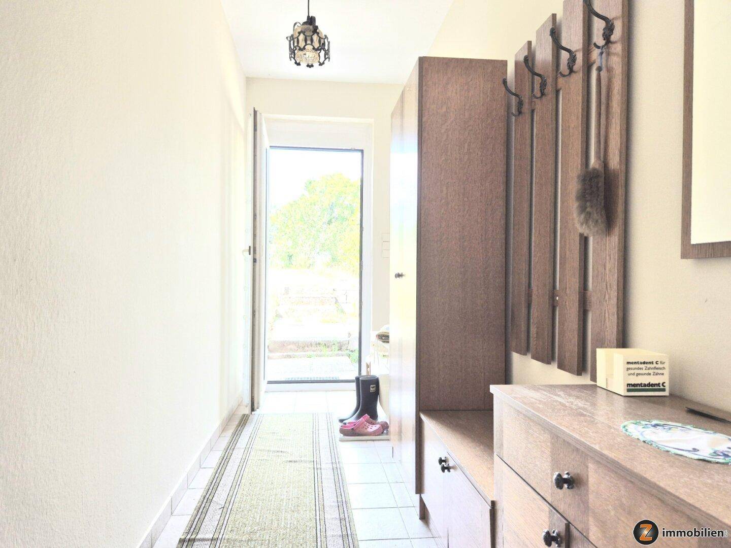 Heller Eingangsbereich mit Fliesenboden, Garderobe und Blick ins Grüne durch die offene Tür.