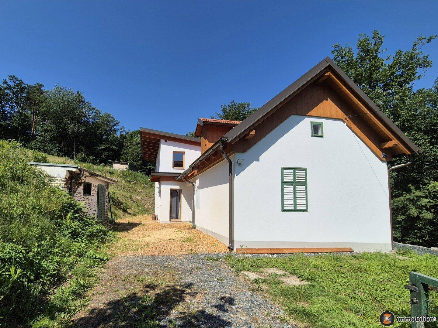 Seitenansicht des Hauses mit weißer Fassade, grünen Fensterläden und Schotterzufahrt umgeben von Natur.
