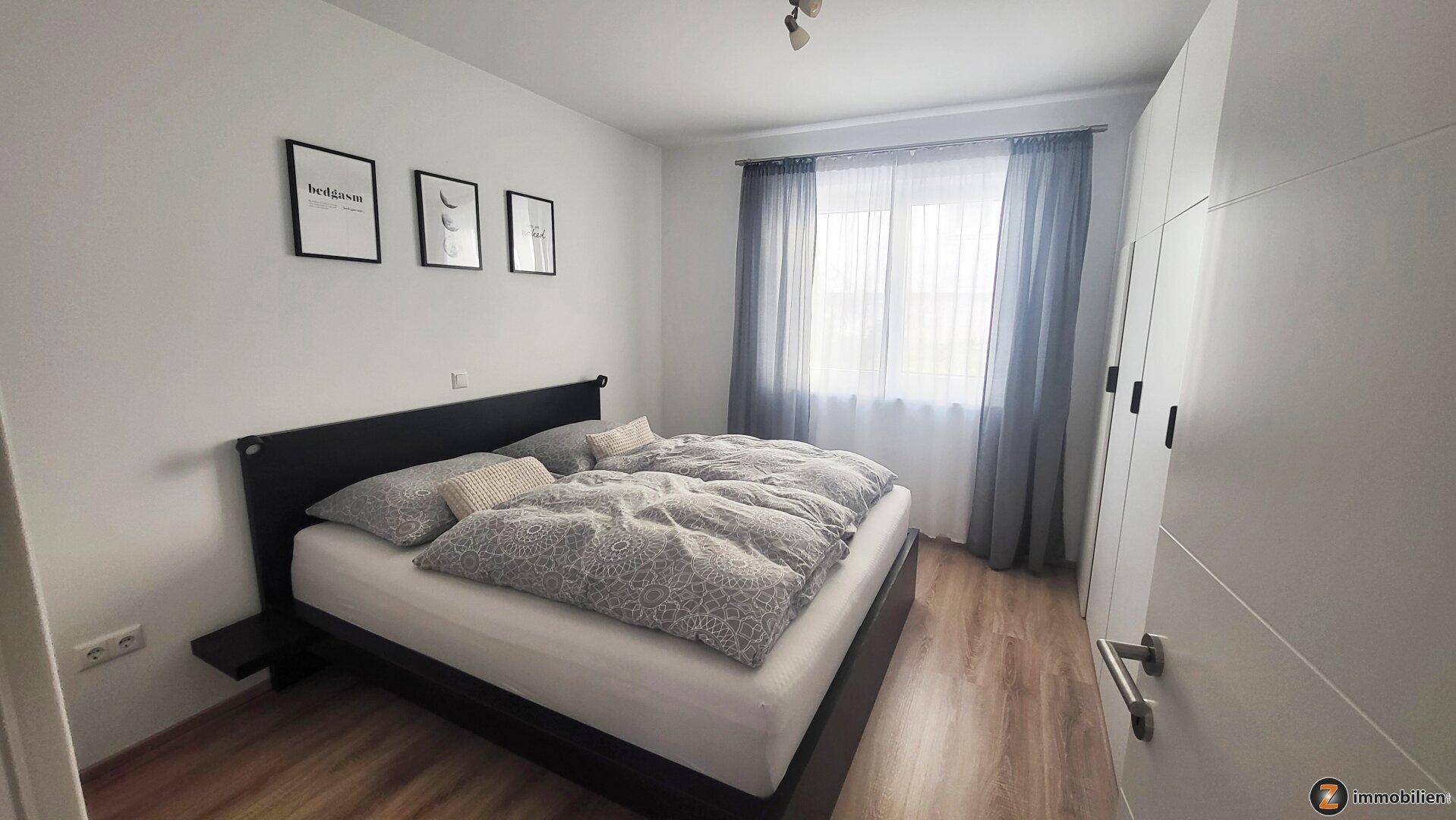 Helles Schlafzimmer mit Doppelbett, Kleiderschrank und Fenster mit Vorhängen.