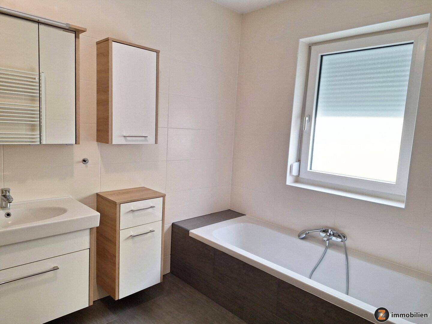 Modernes Badezimmer mit Badewanne, Einzelwaschbecken, Spiegelschrank und Fenster für Tageslicht.