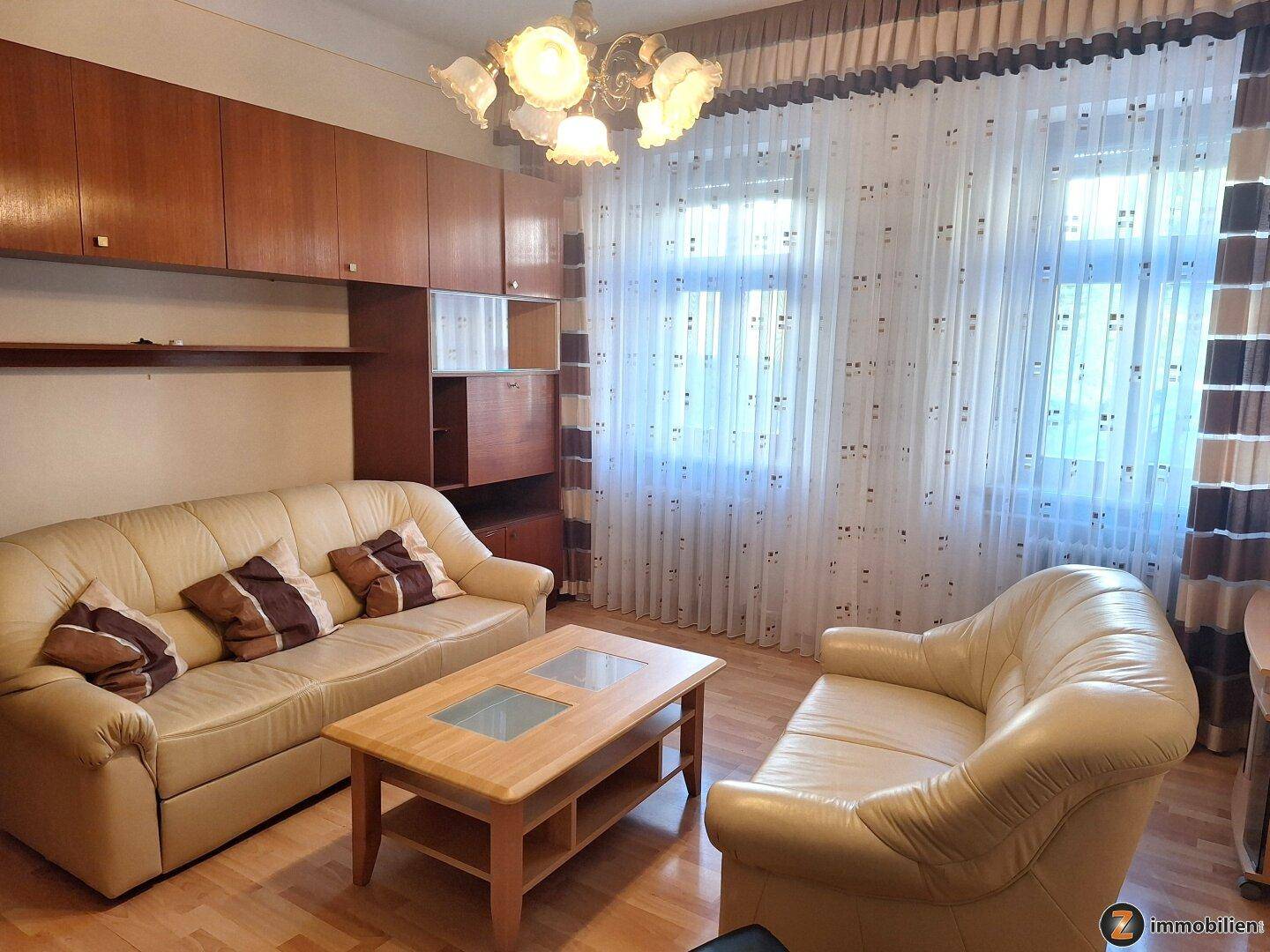 Gemütliches Wohnzimmer mit beigen Sofas, Holzmöbeln und großen Fenstern mit Vorhängen.
