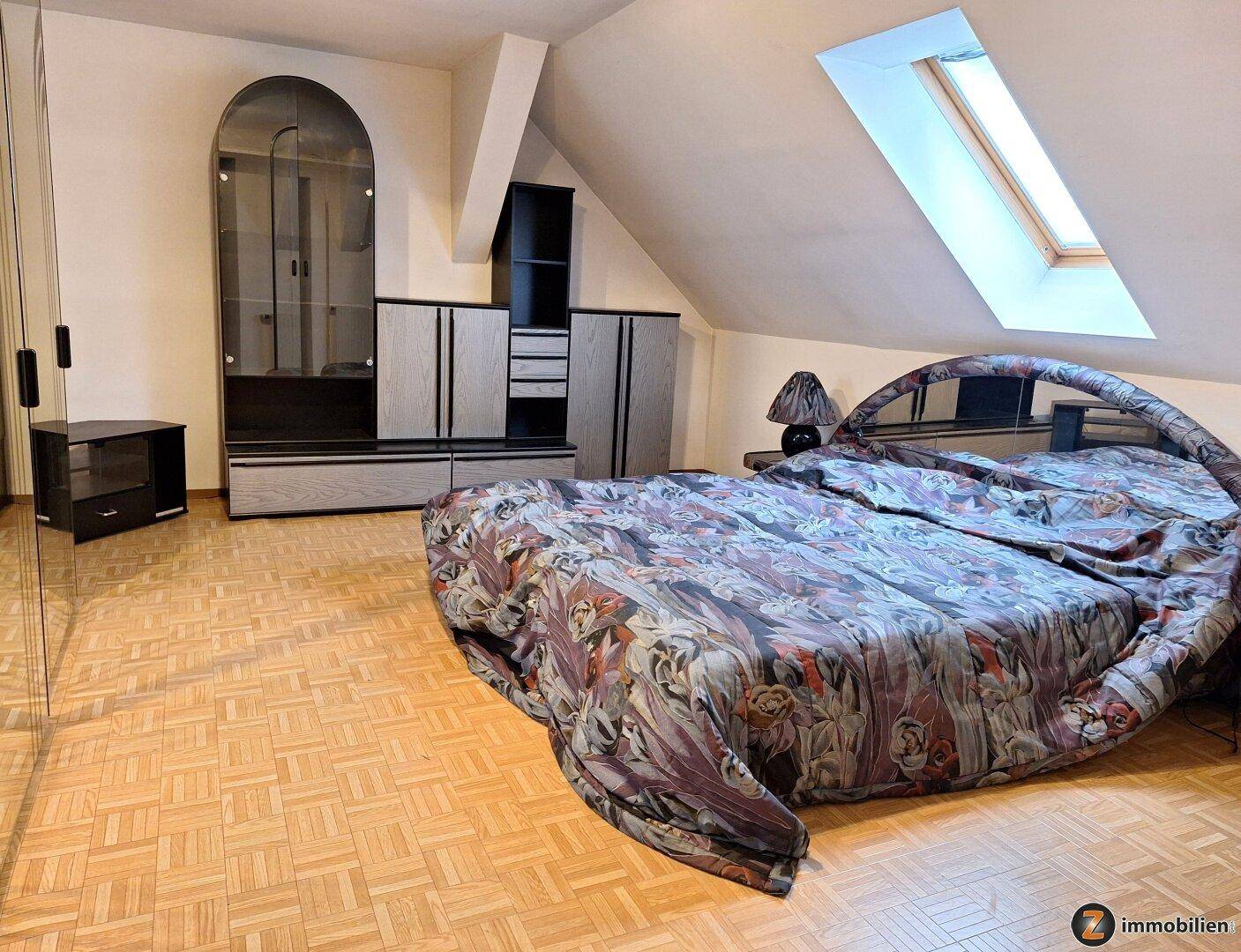 Schlafzimmer im Dachgeschoss mit großem Bett, Parkettboden und einem Dachfenster für natürliches Licht.